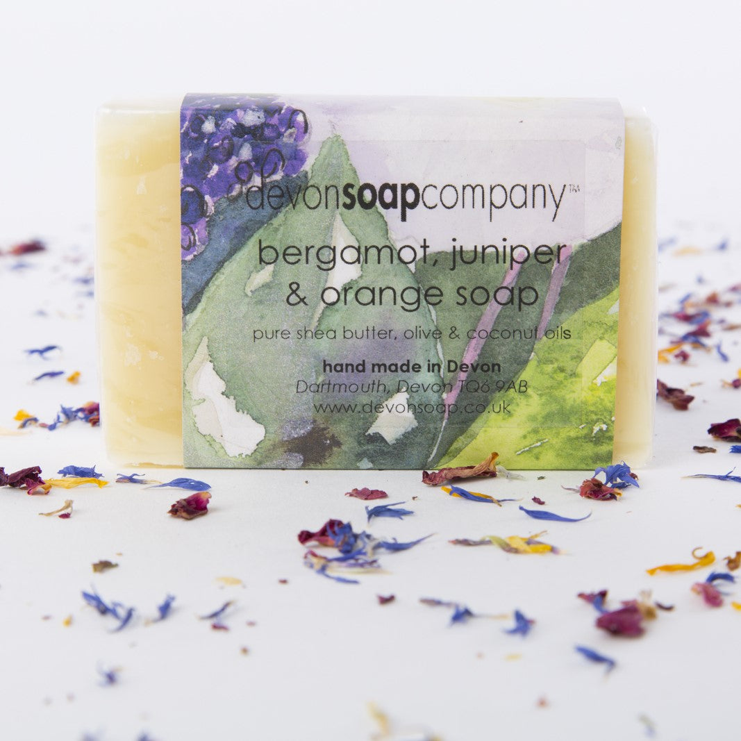 Bergamot, Juniper and Orange soap bar by The Devon Soap Company, wrapped in a watercolour-paper label printed with the words hand made in Devon, Dartmouth, Devon TQ6 9AB, surrounded by scattered dried flower petals