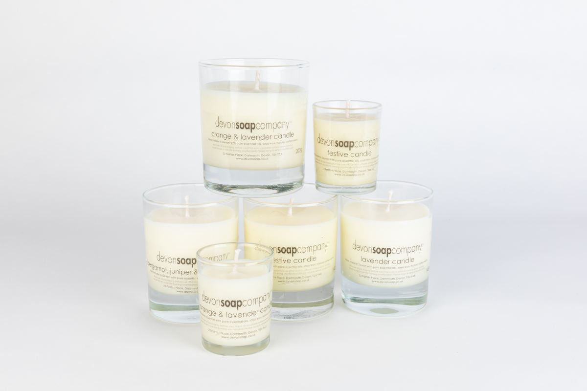 A stacked tier of devonSOAPcompany branded soy-wax candles in clear glass tumblers, each labelled orange and lavender, festive, bergamot juniper and lavender, the Dartmouth address printed on each label